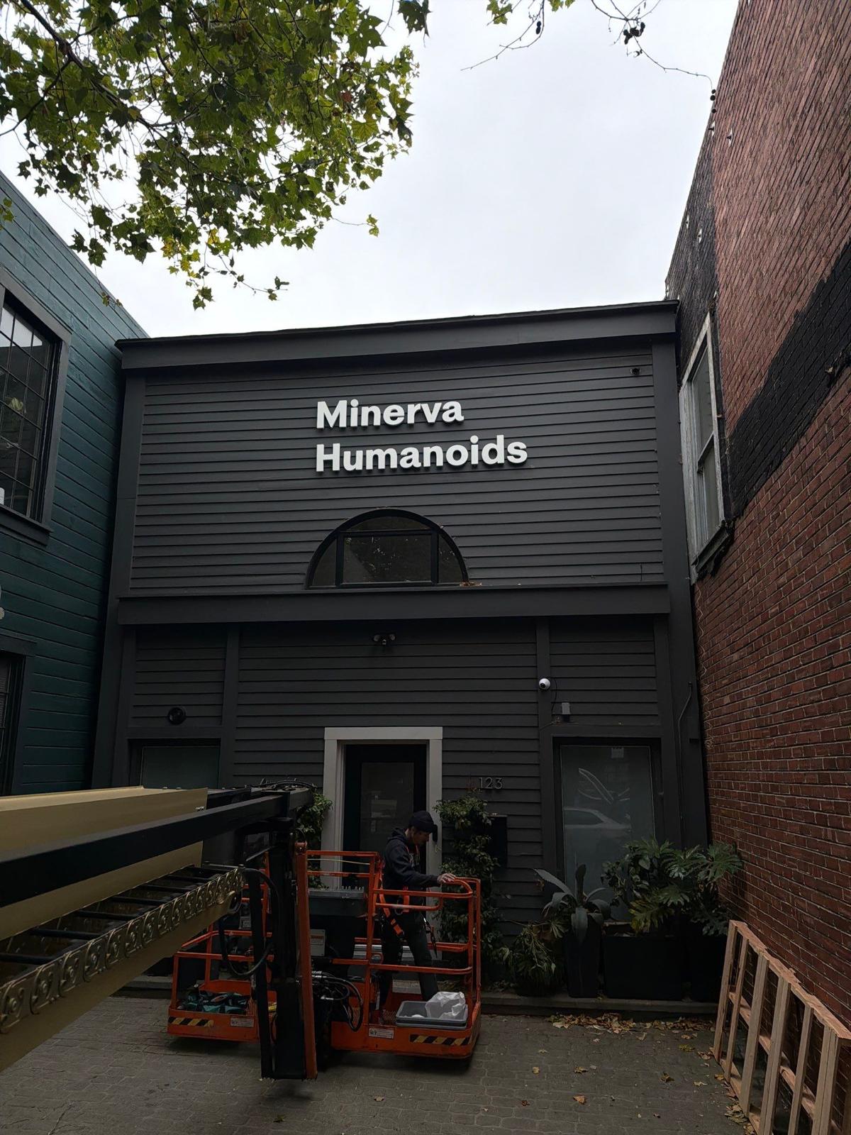 A dark gray building facade with the words 'Minerva Humanoids' in white text above a doorway. An orange cherry picker is parked in front, with a brick wall on the right and another building on the left.