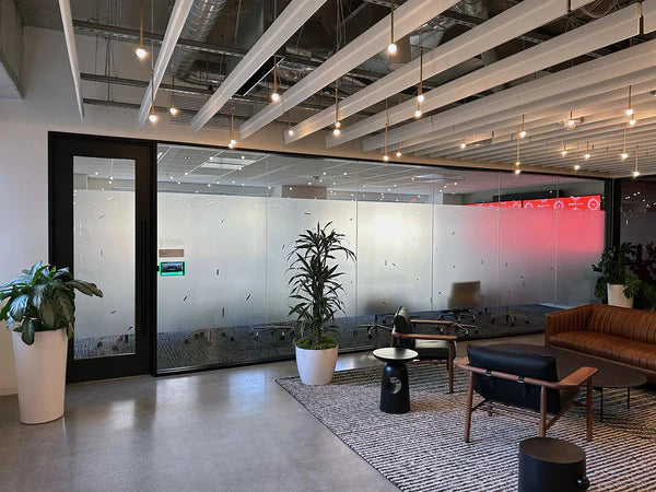 A modern office lobby or lounge area with frosted glass walls, exposed ceiling beams, pendant lights, potted plants, a patterned rug, black and brown chairs, and a brown sofa.