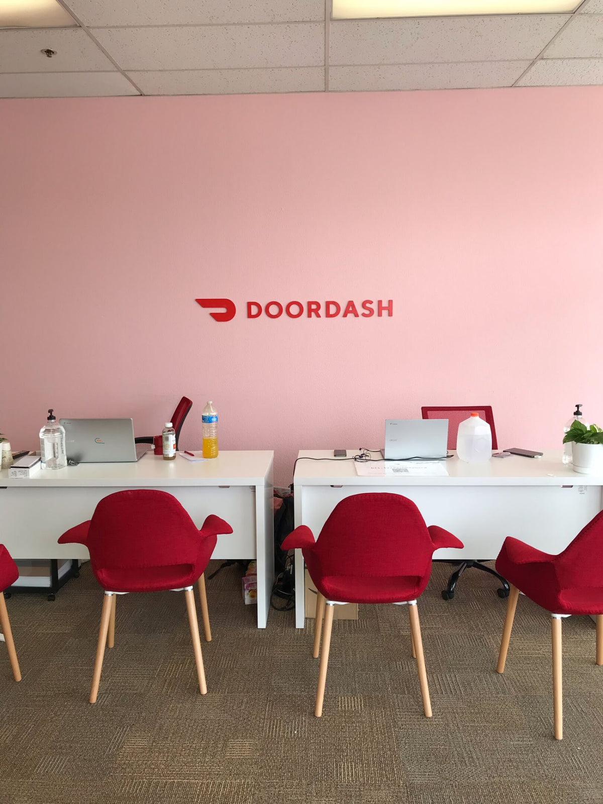 An office interior with a pink wall featuring the red DOORDASH logo, white desks, and red chairs. Several laptops are open on the desks, and the room is viewed from above.
