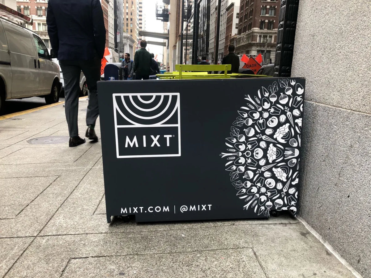 A black outdoor advertising panel with the MIXT logo and white decorative artwork, placed on a city sidewalk next to a stone wall.